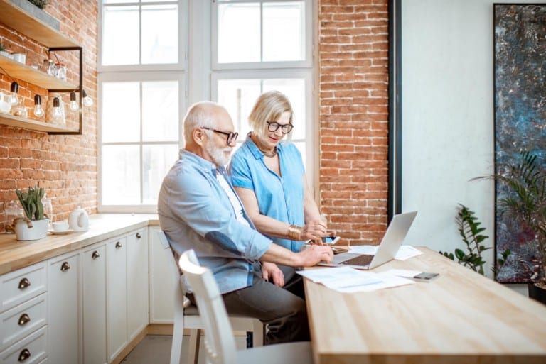 Beautiful senior couple in blue shirts sitting together with laptop on the kitchen at home