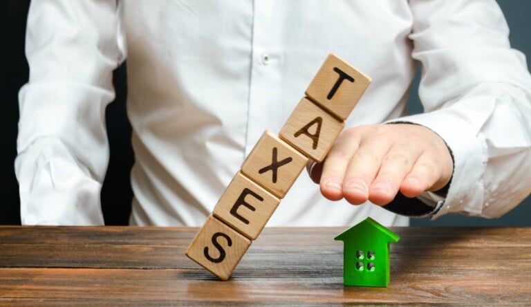 A man protects his hand with a figurine of a house from a falling tower of cubes with the word taxes.